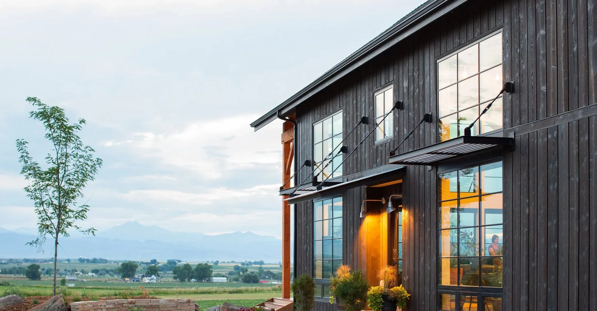 A Frameworks home with dark stained vertical cedar siding and steel awnings, lit from inside at dusk