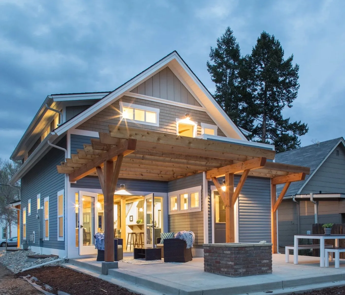 A shingled home with a timber entry porch, lit from inside at dusk
